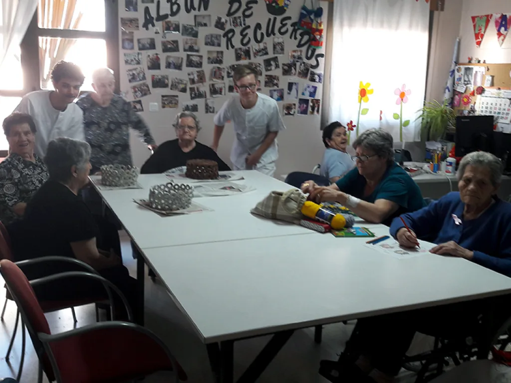 Bewohnerinnen und Bewohner sitzen mit Betreuenden an einem Tisch im Aufenthaltsraum.